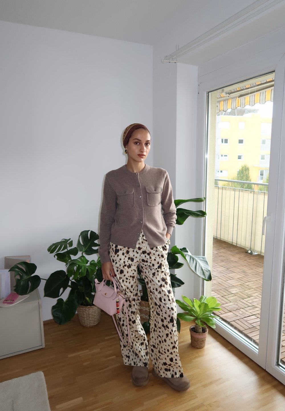 Beige cardigan met twee voorzakken, gecombineerd met wijde geprinte broek in crèmekleur met bruine stippen. Houdt een kleine roze handtas vast.