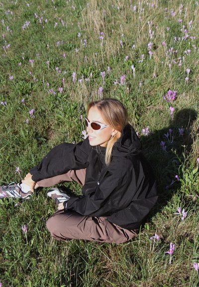 Una chaqueta cortavientos negra con capucha, pantalones cargo marrones y zapatillas deportivas con estampado, sentado en la hierba rodeado de flores moradas.