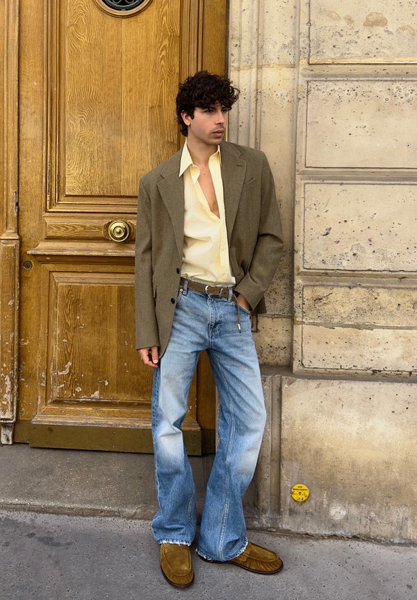 Jonge man met krullend haar die een bruine blazer, lichtbeige overhemd, blauwe spijkerbroek en bruine schoenen draagt, staand bij een houten deur en stenen muur.
