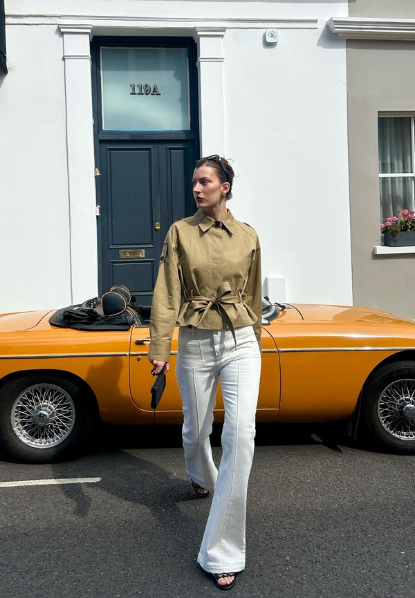 Femme en veste beige et pantalon blanc marchant devant une voiture décapotable rétro orange devant une porte bleu foncé numérotée 119A.