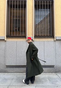 Άνδρας που φορά μακρύ σκούρο παλτό, κόκκινο σκουφάκι, γυαλιά ηλίου και λευκά παπούτσια περπατάει δίπλα από ένα μπεζ και γκρι κτίριο με παράθυρα με κάγκελα.