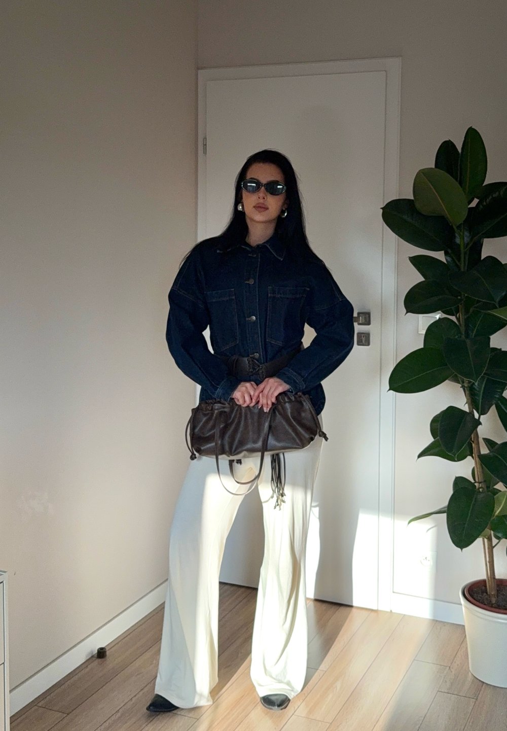 Jachetă din denim cu buzunare mari, strânsă în talie cu un brâu, asortată cu pantaloni albi cu picioare largi și o poșetă din material textil maro.