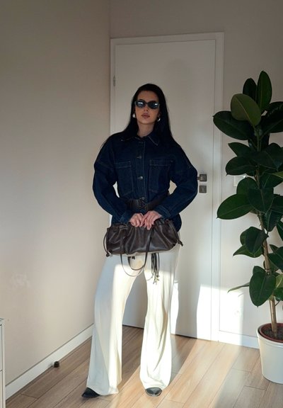 Chaqueta de mezclilla con grandes bolsillos, ceñida a la cintura con un cinturón, combinada con pantalones anchos blancos y un bolso marrón fruncido.