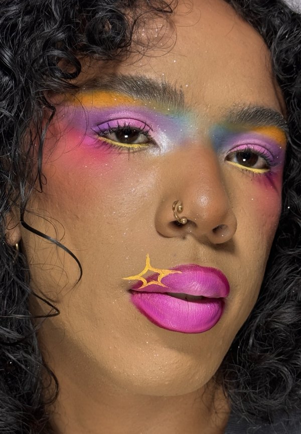 Personne aux cheveux noirs bouclés portant un fard à paupières arc-en-ciel coloré, un rouge à lèvres rose vif avec un détail étoile jaune et un anneau de nez en or.