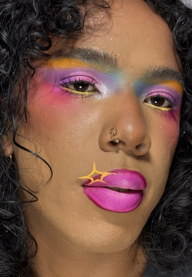 Personne aux cheveux noirs bouclés portant un fard à paupières arc-en-ciel coloré, un rouge à lèvres rose vif avec un détail étoile jaune et un anneau de nez en or.