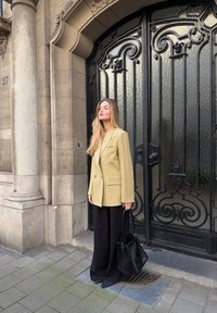 Un blazer beige oversize avec un bouton unique, associé à un pantalon noir à jambes larges et un sac seau noir, devant une grille noire ornée.
