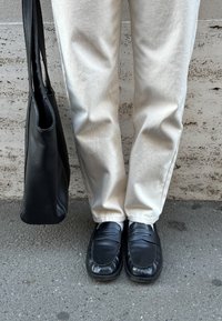 Sorte læder loafers med broderede detaljer, parret med lyse beige bukser. En sort læder tote taske hviler imod benet.