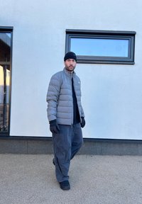 Casaco puffer cinza, luvas pretas, t-shirt preta e calças largas cinzas escuras. De pé numa superfície texturizada junto a janelas num edifício moderno.