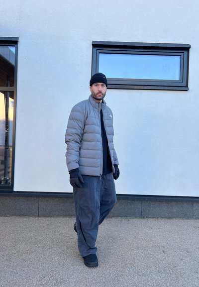 Chaqueta acolchada gris, guantes negros, camiseta negra y pantalones sueltos de gris oscuro. De pie sobre una superficie texturizada junto a ventanas en un edificio moderno.