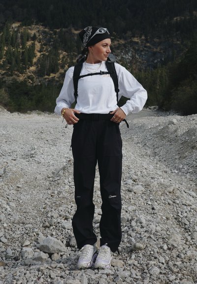 Mujer con ropa de senderismo y una mochila, de pie en un camino rocoso con una montaña boscosa al fondo.