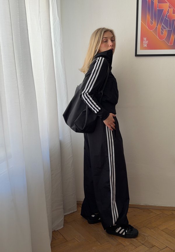 Blonde woman in black Adidas tracksuit with white stripes stands sideways by sheer white curtain, carrying matching black bag indoors.
