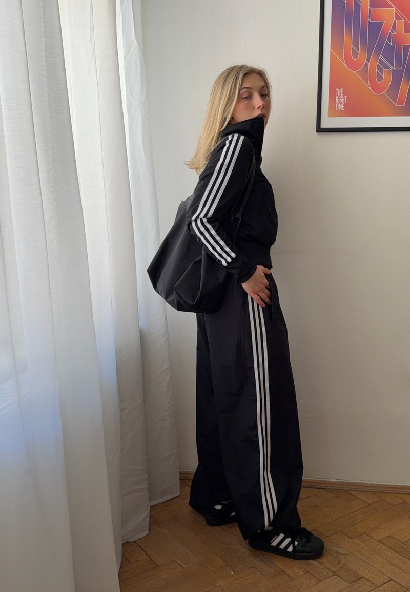 Femme blonde en survêtement Adidas noir avec des bandes blanches, debout de profil près d'un rideau blanc transparent, portant un sac noir assorti à l'intérieur.