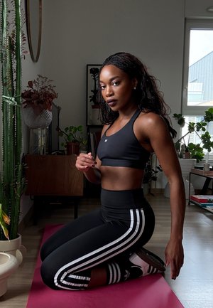 Top deportivo corto negro y leggings de cintura alta con rayas blancas a los lados. Sentada sobre una esterilla rosa, rodeada de plantas de interior.