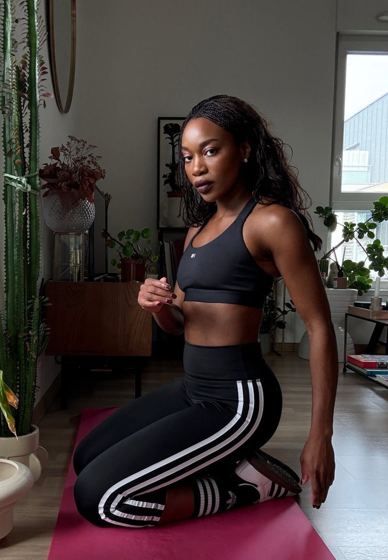 Fekete atlétikai crop top és magas derekú leggings fehér oldalsó csíkokkal. Rózsaszín matracon ül, beltéri növényekkel körülvéve.