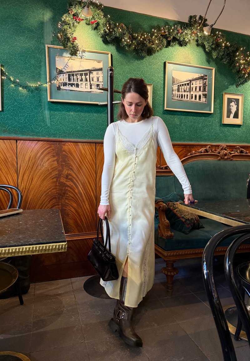 Kvinna som bär en creamfärgad slipklänning över vit topp med långa ärmar, bruna stövlar, håller en svart handväska, stående i ett dekorerat café i vintage-stil.