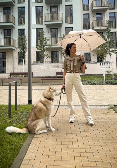 Un husky de color marrón claro y blanco está sentado al lado de una persona que sostiene un paraguas beige. La persona lleva puesta una blusa de punto, pantalones blancos y zapatillas.