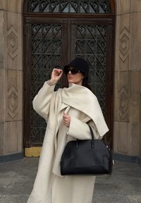 Prevelik beli volneni plašč, črni klobuk v obliki vedra in velika črna usnjena torba. Stoji ob izrezljanih lesenih vratih z geometrijskimi vzorci.