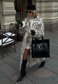 Fuskepelskåpe med et prikkete mønster, hvit høyhalset genser, høye svarte støvler, svarte hansker og en strukturert svart håndveske, holder en kaffekopp.