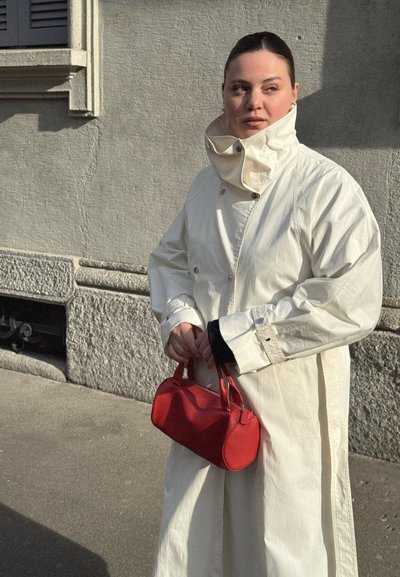 Mujer de cabello oscuro con un abrigo blanco de cuello alto sostiene un pequeño bolso rojo, de pie contra una pared de piedra texturizada iluminada por el sol.