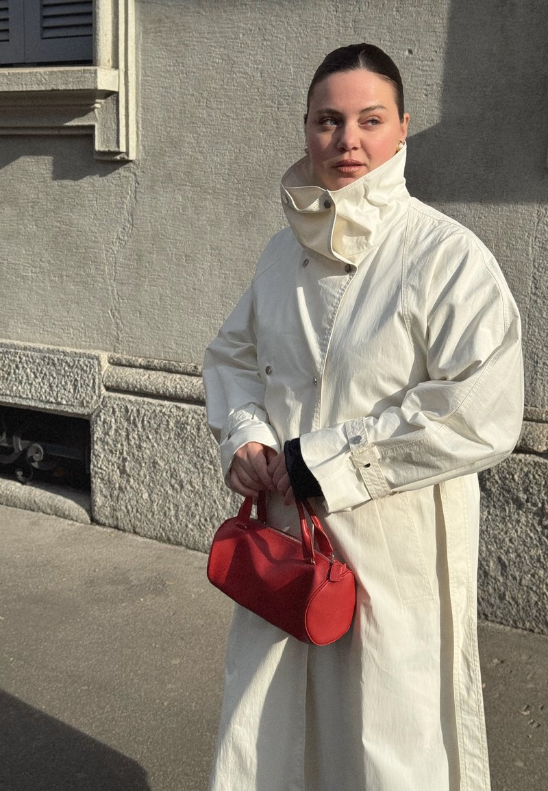 Mujer de cabello oscuro con un abrigo blanco de cuello alto sostiene un pequeño bolso rojo, de pie contra una pared de piedra texturizada iluminada por el sol.