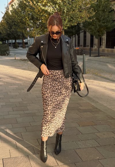 Chaqueta de cuero negra, blusa negra ajustada y una falda midi de estampado de leopardo. Botines negros y un pequeño bolso negro. Entorno exterior pavimentado.