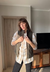 Femme à l'intérieur portant une chemise à carreaux beige et une veste zippée vert foncé, ajustant le col de la veste, debout près d'une porte en bois et d'un meuble TV.