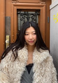 Ung kvinde med langt sort hår iført en fluffy beige frakke og mørk top, stående foran en brun dør med dekorativt jernarbejde.