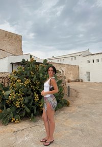 Top bianco senza maniche e pantaloni corti decorati con orlo frangiato. Infradito nere. Lo sfondo presenta un cactus con frutti gialli e muri rustici.