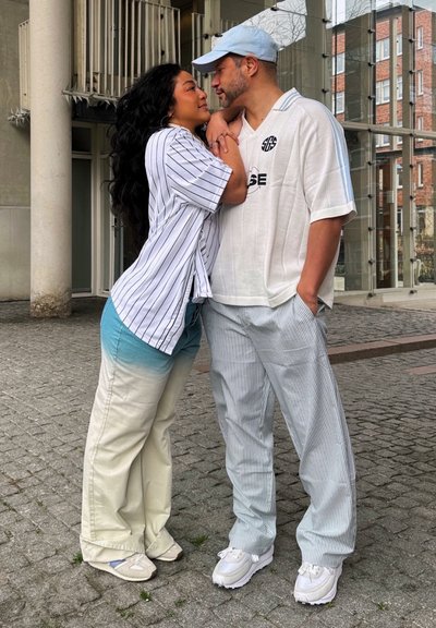 Dos personas están de pie cerca, vistiendo atuendos informales. Uno lleva una camisa a rayas blanca y azul con pantalones claros en ombre. El otro lleva una camisa blanca lisa y pantalones a rayas.