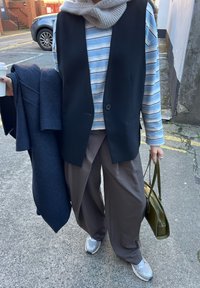 Černá vesta bez rukávů přes pruhovanou košili s dlouhým rukávem, šedé široké kalhoty a šedé tenisky. Drží tmavý kabát a zelenou kabelku.