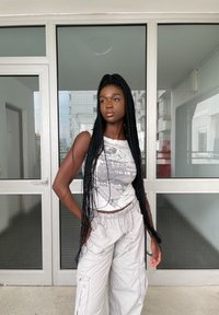 Λευκή γραφική μπλουζά με χειροποίητο σχέδιο; ανοιχτό γκρι cargo παντελόνι με πλαϊνές τσέπες. Μακριά πλεγμένα μαλλιά, στέκεται μπροστά από γυάλινες πόρτες.