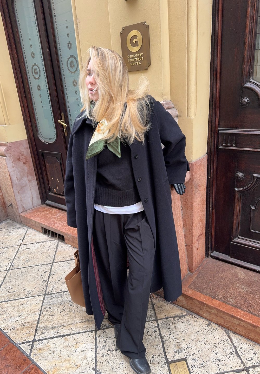 Melnas vilnas mētelis ar lielām pogām, uzlikts virs melna džemperi un baltas krekla; apvienots ar platām melnām biksēm un zaļu šalli. Brūna rokassoma.