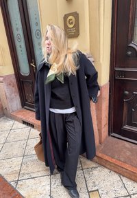 Melnas vilnas mētelis ar lielām pogām, uzlikts virs melna džemperi un baltas krekla; apvienots ar platām melnām biksēm un zaļu šalli. Brūna rokassoma.