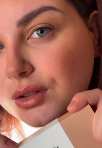 Lähikuva henkilöstä, joka pitää käsissään tuotepakkausta; ominaisuuksiin kuuluvat sileä iho, kiiltävät luonnolliset huulet ja kevyt silmämeikki, pehmeä valaistus ja neutraalit sävyt.