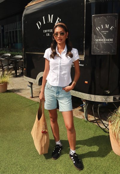 Mujer con gafas de sol, camiseta blanca de manga corta, pantalones cortos de mezclilla y zapatillas negras, sosteniendo un gran bolso beige, de pie cerca de un camión negro de café.