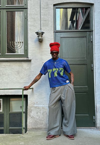 Jersey de malla azul con impresión verde '777', pantalones grises holgados, gorro de punto rojo y gafas de sol negras, con una barandilla verde y una puerta al fondo.