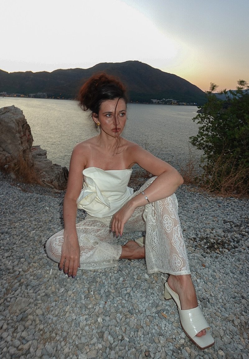 Weisses drapiertes Oberteil, Spitzen-Hose mit weitem Bein und Blumenmuster sowie weisse Sandalen mit Blockabsatz. Kieshintergrund, Wasser und Berge sichtbar.