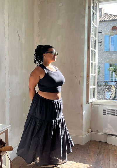 Top corto negro y una larga falda negra con volantes. La luz natural que entra por la ventana ilumina la pared texturizada y el suelo de madera.