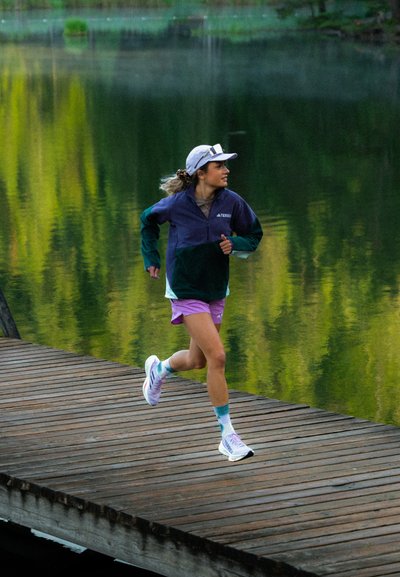 Ropa de correr que incluye una chaqueta verde oscuro y morada, pantalones cortos morados claros, calcetines blancos y zapatillas de correr multicolores sobre un muelle de madera.
