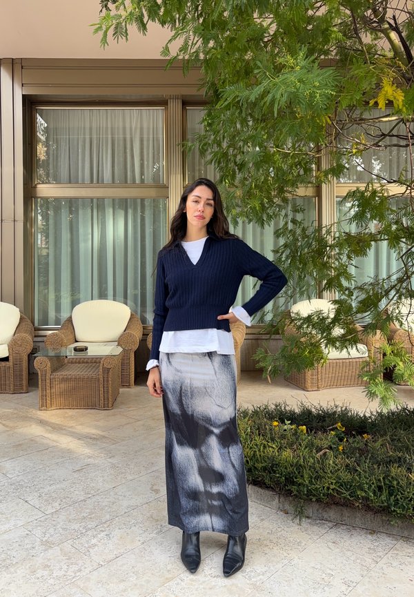 Žena stojaca na terase vedľa prútených stoličiek a skleneného stola, oblečená v čiernom svetri, dlhá čierno-biela sukňa a čierne členkové čižmy.