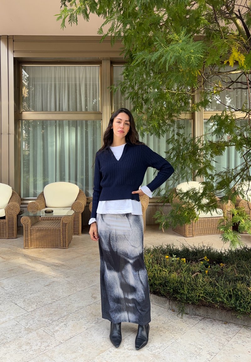 Donna in piedi sul patio accanto a sedie in vimini e tavolo in vetro, indossa un maglione nero, una gonna lunga bianca e nera e stivali neri alla caviglia.