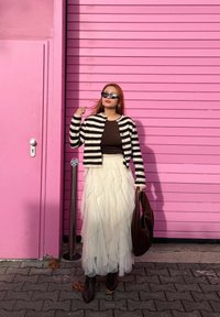 Cardigan rayé noir et blanc porté sur un haut marron, associé à une jupe en tulle blanche. Sac marron et bottes foncées, sur un fond rose.