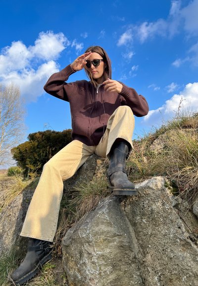 Sudadera marrón con cremallera, pantalones anchos beige, botas negras hasta la rodilla, sentado en una roca con un cielo azul y nubes de fondo.