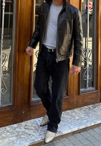 Személy fekete bőrdzsekit, szürke pólót, fekete farmert és kígyómintás hegyes orrú csizmákat viselve áll egy márványpadlós küszöbön, egy fa ajtónál.