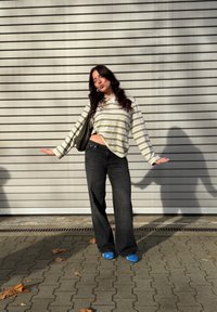 Tričká s dlhým rukávom s pruhmi a bielym golierom, čierne rifle s vysokým pásom a modré topánky, stojace oproti sivému múru s líniami.