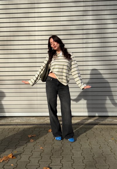 Camisa de manga larga a rayas con cuello blanco, jeans negros de tiro alto y zapatos azules, de pie contra una pared gris con líneas.