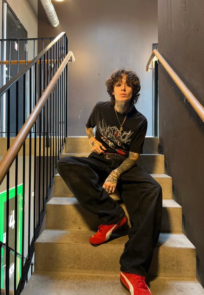 Camiseta gráfica negra con diseño rojo y blanco, jeans oversized negros y zapatillas rojas; sentado en escalones de concreto con una barandilla de metal.