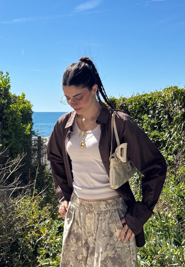 Femme debout près de la verdure au bord de la mer, portant des lunettes de soleil, une chemise sombre ouverte, un débardeur blanc, un pantalon à motifs et un petit sac sur l'épaule.