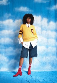 Yellow knitted v-neck sweater with maroon stripes, white shirt underneath, dark denim shorts, red heeled boots, and striped socks set against a blue background.