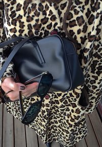 Sac à main en cuir noir à texture lisse, forme arrondie, et fermeture éclair, accompagné de lunettes de soleil foncées avec un motif décoratif.